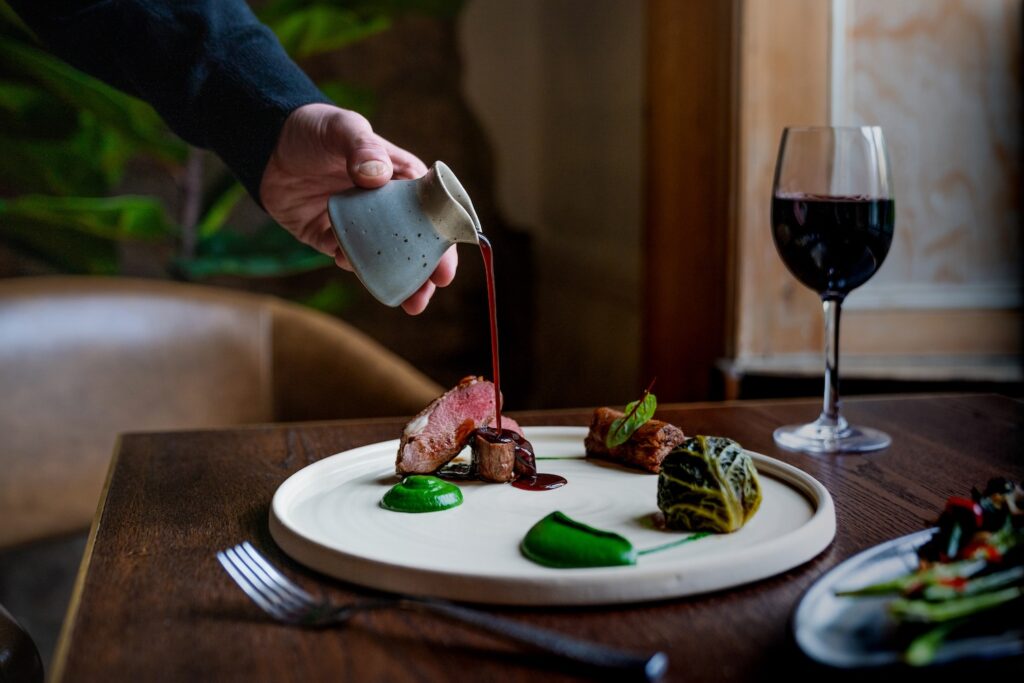 Person pouring a rich red wine jus over a plate of second spring lamb, or hogget, with a glass of red wine