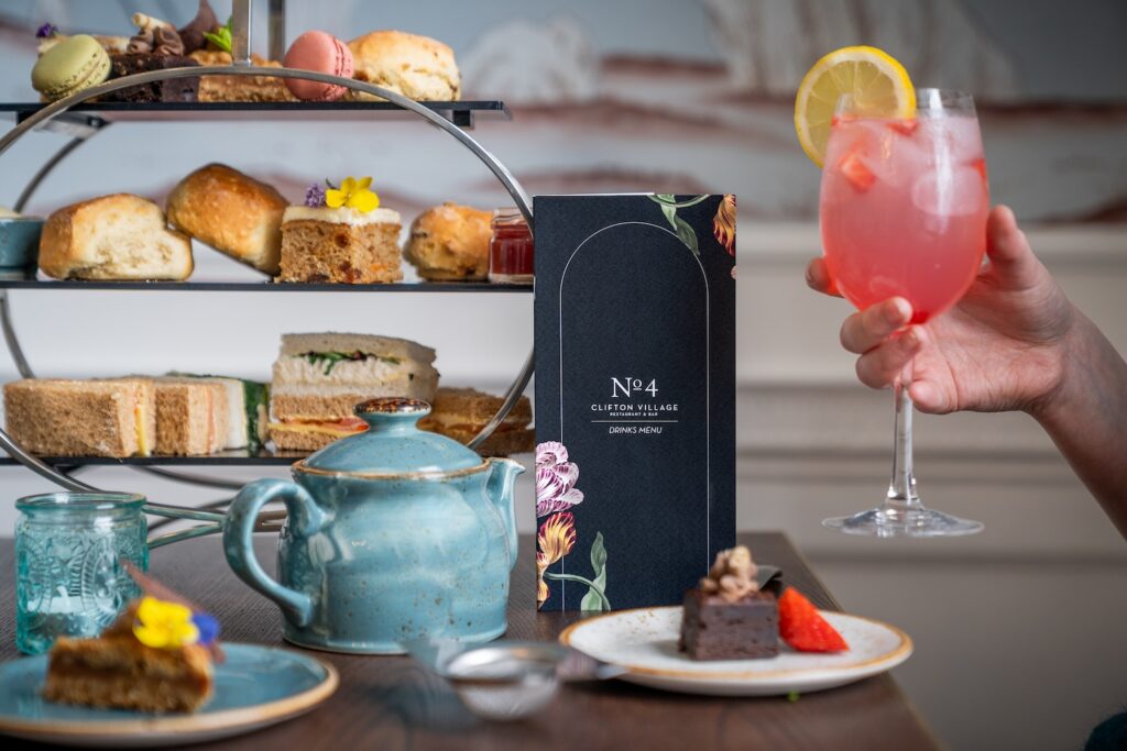 An elegant afternoon tea setup featuring a variety of sandwiches and pastries, with a person holding a refreshing pink cocktail