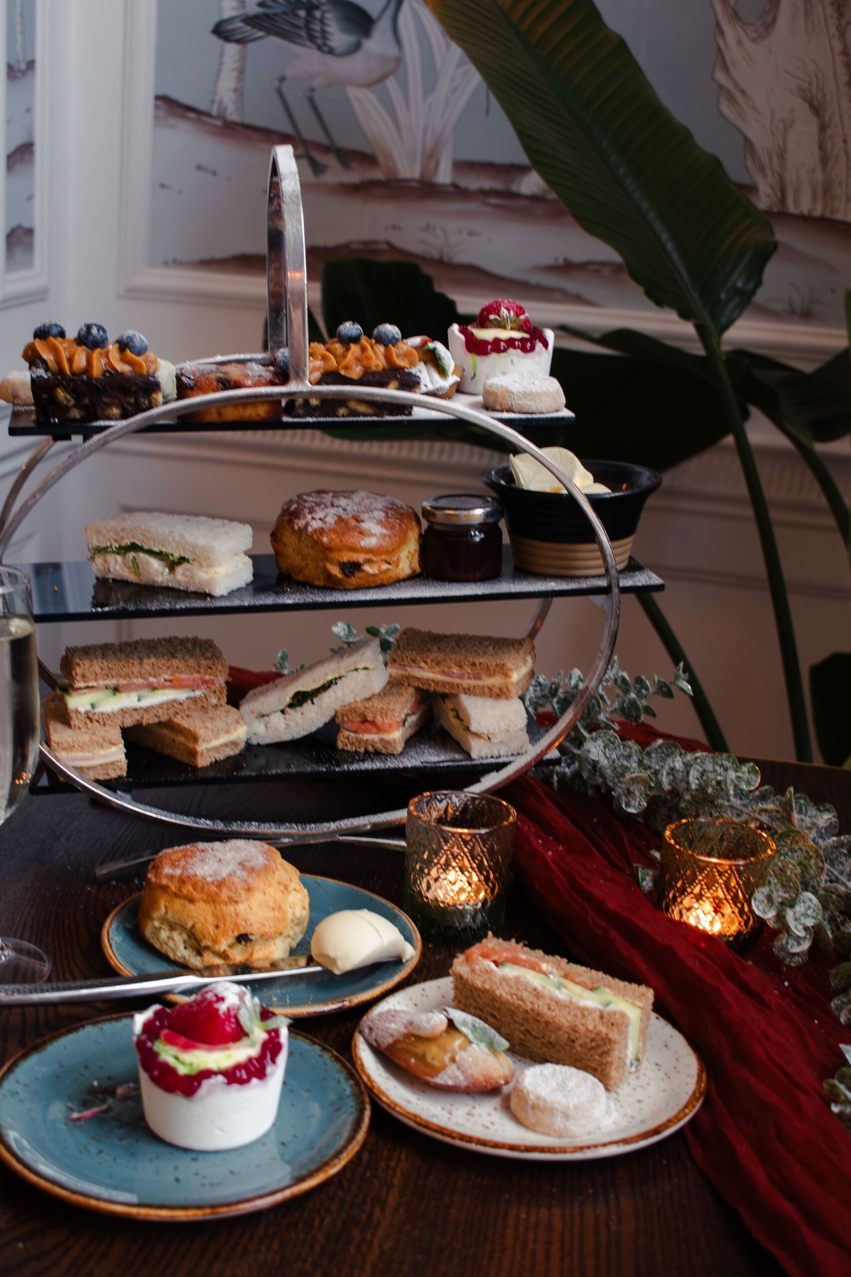 Elegant winter afternoon tea at No.4 Clifton Village in Bristol, featuring a festive tiered stand with finger sandwiches, raisin scones with clotted cream and jam, and decadent patisserie topped with fresh berries. A glass of prosecco and flickering candles add warmth to the Christmas-themed table setting with frosted greenery and red fabric accents.